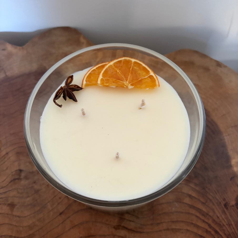Candle in a glass jar with an orange slice and star anise on top, placed on a wooden surface.